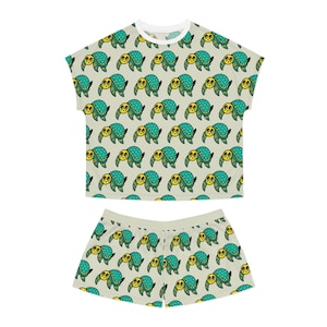 May include: A short-sleeve t-shirt and shorts set in a light beige color featuring an all-over repeating pattern of cartoon turtles with teal shells, yellow faces, and dark outlines. The set includes a crew neck top and matching elastic waist shorts.