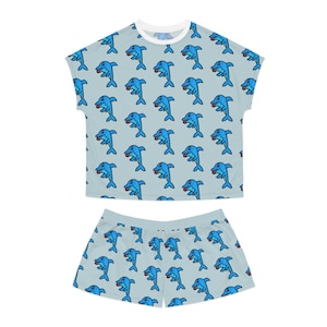May include: A light blue pajama set consisting of a short-sleeved top and matching shorts. The fabric features an all-over repeating pattern of cartoon dolphins in blue with black outlines, each depicted in a playful pose on a light blue background.