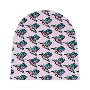 May include: A light pink beanie featuring a repeating pattern of stylized hummingbirds. Each hummingbird is outlined in black and filled with a gradient of vibrant colors including blue, green, and pink, creating a consistent graphic design.