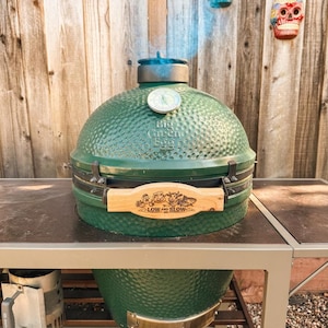 May include: A green ceramic Big Green Egg grill with a wooden handle that reads "LOW and SLOW". The grill is on a metal stand. Decorative skulls hang in the background.