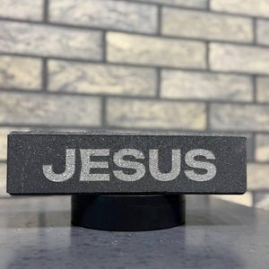 Pode incluir: Um bloco retangular cinza escuro com a palavra "JESUS" em letras prateadas brilhantes. O bloco está sobre uma base preta circular, com uma parede de tijolos desfocada ao fundo.