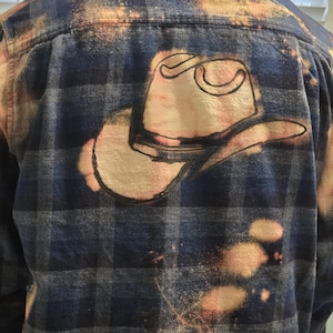 May include: Blue and black plaid flannel shirt with a cowboy hat design on the back. The hat is outlined in black and has a light beige fill. The shirt has a distressed, bleached effect.