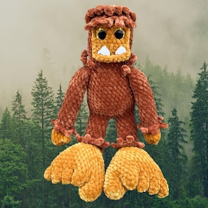May include: A handmade plush Bigfoot doll in shades of brown and tan. The doll has large feet, a square head, and small black eyes. The background features a forest scene with tall green trees and a misty sky.