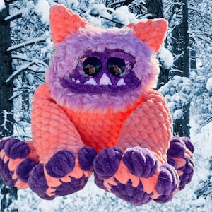 May include: A handmade plush monster toy with a vibrant color scheme. The monster features a fuzzy purple head and ears, a smiling purple mouth, and large, reflective eyes. Its body is a textured orange, and its feet are purple. The background is a snowy forest.