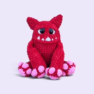 May include: A plush monster toy crafted from deep red yarn with a textured, fuzzy appearance. It features large, glossy black eyes, a wide mouth with visible white teeth, pointed ears, and oversized feet with distinct light pink circular pads.