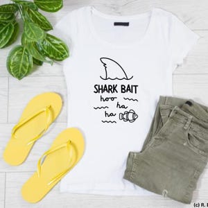 May include: A white t-shirt with the text "SHARK BAIT" and a shark fin graphic. The image also includes yellow flip-flops, green pants, and a green plant on a light wood-grain surface.