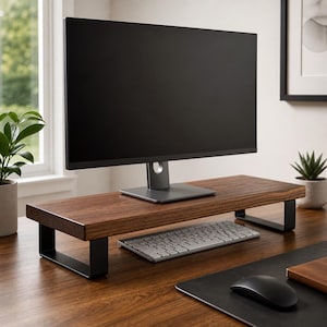 May include: A dark brown wooden monitor stand with black metal legs. A computer monitor sits on the stand, with a wireless keyboard and mouse below. The stand is on a wooden desk, with a plant in the background.