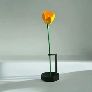 Könnte beinhalten: Eine gelbe Tulpenblume mit grünem Stiel in einer schwarzen geometrischen Vase. Die Tulpe hat leuchtend gelbe Blütenblätter und einen grünen Stiel. Die Vase ist modern, mit sechseckiger Basis und rechteckiger Halterung. Die Vase ist etwa 15 cm hoch.