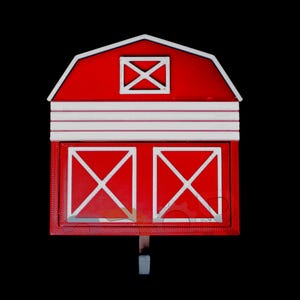 May include: A red barn-shaped decorative item with white trim and accents. The barn features a white cross design on the doors and a small window. It has a hook for hanging and is set against a black background.