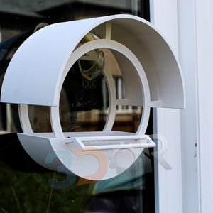 Peut inclure: Un mangeoire à oiseaux blanc de forme circulaire, avec un toit incurvé. Il est conçu pour être fixé à une fenêtre. Le design est minimaliste et moderne, avec un petit perchoir. Dimensions: environ 30 cm de diamètre.