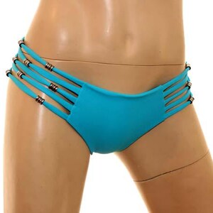 May include: A turquoise bikini bottom with multiple thin straps on each side, adorned with small silver-colored metal accents. The swimwear is displayed against a white background, showcasing its design and color.
