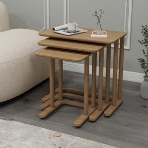 Puede incluir: Un juego de tres mesas nido de roble claro. Las mesas son rectangulares con esquinas redondeadas y de diferentes alturas. Una taza y un periódico enrollado están sobre la mesa superior. Las mesas están en un salón.