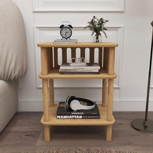 May include: A three-tiered wooden side table with a natural finish. The top shelf holds a black alarm clock, books, and a small vase with greenery. The middle shelf displays books with the letters "CMD" visible. The bottom shelf has headphones and a book titled "MANHATTAN AN ISLAND IN FOCUS."