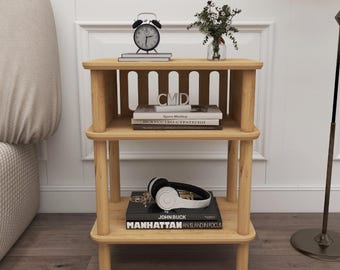 May include: A three-tiered wooden side table with a natural finish. The top shelf holds a black alarm clock, books, and a small vase with greenery. The middle shelf displays books with the letters "CMD" visible. The bottom shelf has headphones and a book titled "MANHATTAN AN ISLAND IN FOCUS."