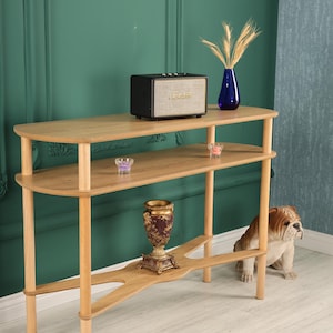 May include: A light wood console table with three shelves. A black speaker, blue vase with wheat, and decorative items are displayed on the shelves. A bulldog statue sits nearby. The table is 48 inches long, 12 inches wide, and 30 inches tall.