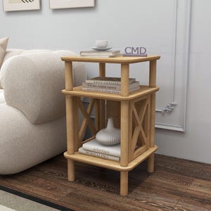 May include: A three-tiered, light-colored wooden side table with a square top and bottom. The table has a geometric design on the sides and holds books, a vase, and a teacup. The letters "CMD" are visible on the top.