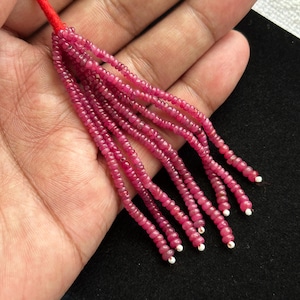 Puede incluir: Una borla compuesta por múltiples hilos de pequeñas cuentas redondas de color rosa intenso. Los hilos están unidos en la parte superior por un cordón rojo brillante y cada hilo termina con una pequeña cuenta blanca en el extremo inferior.