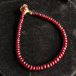 Peut inclure: Un bracelet composé de petites pierres précieuses rondes et rouge foncé. Le bracelet est enfilé et possède un fermoir doré. Les pierres précieuses sont d'un rouge foncé riche, et le bracelet est présenté sur un fond noir.
