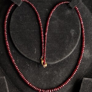 Peut inclure: Un collier composé de petites perles rondes, rouge foncé. Les perles sont enfilées ensemble, formant un brin continu. Le collier est présenté sur une surface noire, avec un fermoir doré.