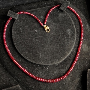 Puede incluir: Collar de cuentas de rubíes rojos, compuesto por pequeñas cuentas redondas. El collar se muestra sobre un fondo negro de terciopelo. El cierre dorado añade un toque de contraste. Una joya elegante y atemporal.
