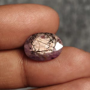 Peut inclure: Une pierre précieuse de taille ovale avec une teinte translucide violet clair et rose, présentant des inclusions sombres et ramifiées en forme d'aiguilles. La surface à facettes reflète la lumière, soulignant les motifs et les nuances internes.
