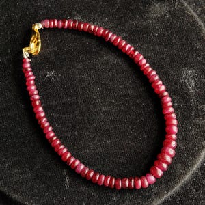 Peut inclure: Un bracelet de perles de rubis rouges foncés, rondes et de petite taille, avec un fermoir doré. Le fermoir est de couleur or vif, contrastant avec le rouge foncé des perles. Le bracelet est présenté sur une surface noire.
