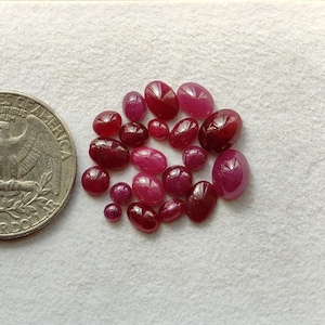 May include: A collection of small, oval-shaped, polished red rubies in varying shades of deep crimson and pinkish-red, arranged in a cluster next to a silver-colored United States quarter coin for a clear visual size reference.