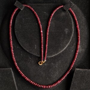 Op de afbeelding: Een robijnen ketting, gepresenteerd op een zwarte ondergrond. De ketting bestaat uit kleine, ronde, dieprode edelstenen die aan elkaar zijn geregen. De sluiting is goudkleurig. De ketting wordt gepresenteerd in een sieradendoos.