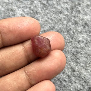Può includere: Una piccola gemma di rubino grezza e non tagliata, di colore rosso intenso e con una forma naturalmente irregolare e sfaccettata. La pietra presenta una superficie ruvida e opaca con sottili variazioni di tono e chiarezza.