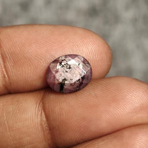 Può includere: Pietra preziosa ovale e sfaccettata con una tonalità rosa-viola. La gemma presenta inclusioni interne e una superficie lucida, che riflette la luce. La pietra è tenuta in una mano, mostrando le sue dimensioni e il suo colore. La gemma è probabilmente un rubino.