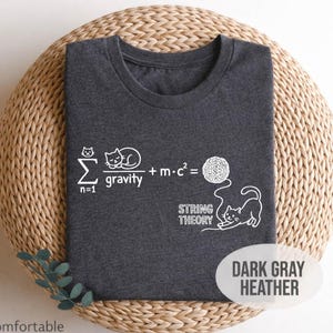 May include: Dark gray heather t-shirt with a white graphic featuring a cat-themed string theory equation. The design includes a sleeping cat, a ball of yarn, and a playful cat. The shirt is displayed on a woven, circular surface.