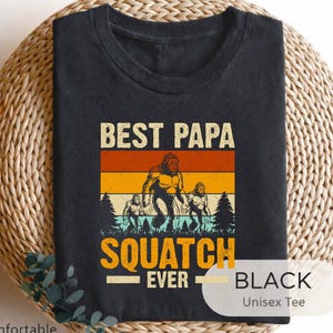 May include: Black unisex tee with a retro design featuring a Bigfoot illustration and the text "BEST PAPA SQUATCH EVER." The design includes a Bigfoot family in a forest setting with orange, yellow, and green stripes. The shirt is labeled "Soft & Comfortable."