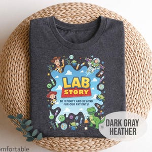 May include: Dark gray heather t-shirt with a colorful graphic featuring characters and the text "LAB STORY TO INFINITY AND BEYOND FOR OUR PATIENTS!". The shirt is labeled "Soft & Comfortable".