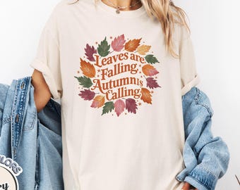 May include: Ivory-colored t-shirt with the text "Leaves are Falling, Autumn is Calling" surrounded by a wreath of colorful leaves. The shirt is part of the Comfort Colors collection.