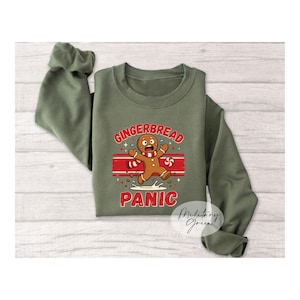May include: An olive green long-sleeve sweatshirt featuring a graphic of a running gingerbread person with a red and white striped scarf. The text reads GINGERBREAD PANIC in bold red letters, with a small peppermint candy and white splash details.