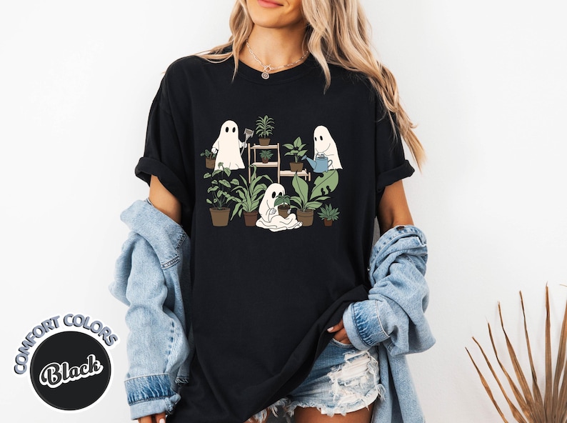 May include: Black t-shirt featuring a whimsical graphic of three cartoon ghosts tending to potted plants. One ghost waters plants, another tends to a shelf, and the third sits with a plant. The shirt has the words "Comfort Colors" and "Black" on it.