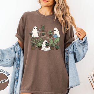 May include: Espresso-colored t-shirt featuring a whimsical graphic of three cartoon ghosts tending to potted plants. The design includes a watering can and a plant stand. The shirt is part of the Comfort Colors collection.