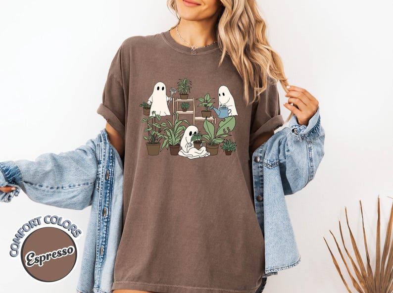 May include: Espresso-colored t-shirt featuring a whimsical graphic of three cartoon ghosts tending to potted plants. The design includes a watering can and a plant stand. The shirt is part of the Comfort Colors collection.