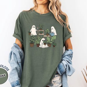 May include: Moss green t-shirt featuring a graphic of three cartoon ghosts tending to plants. One ghost waters plants, another tends to a pot, and the third stands near a shelf of plants. The shirt has rolled-up sleeves.