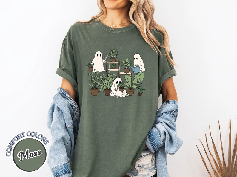 May include: Moss green t-shirt featuring a graphic of three cartoon ghosts tending to plants. One ghost waters plants, another tends to a pot, and the third stands near a shelf of plants. The shirt has rolled-up sleeves.
