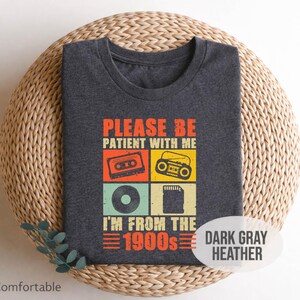 May include: Dark gray heather t-shirt with the text "PLEASE BE PATIENT WITH ME I'M FROM THE 1900s" in orange and white. The design includes retro illustrations of a cassette tape, radio, vinyl record, and a memory card.