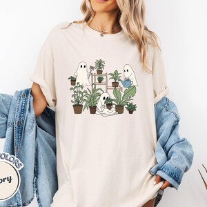 May include: Ivory-colored t-shirt featuring a whimsical illustration of three ghost figures tending to potted plants. The ghosts are depicted with simple, friendly faces, surrounded by various green plants in brown pots. The shirt has the words "Comfort Colors" and "Ivory" on it.