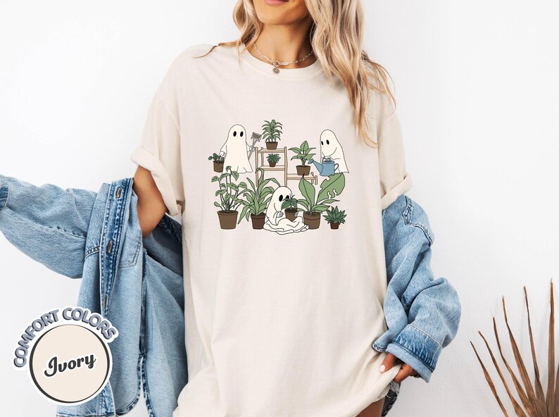 May include: Ivory-colored t-shirt featuring a whimsical illustration of three ghost figures tending to potted plants. The ghosts are depicted with simple, friendly faces, surrounded by various green plants in brown pots. The shirt has the words "Comfort Colors" and "Ivory" on it.