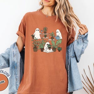 May include: Terracotta-colored t-shirt featuring a graphic of three cartoon ghosts tending to potted plants. The ghosts are white with black eyes, and the plants are green. The shirt has the words "COMFORT COLORS" and "Yam" on it.