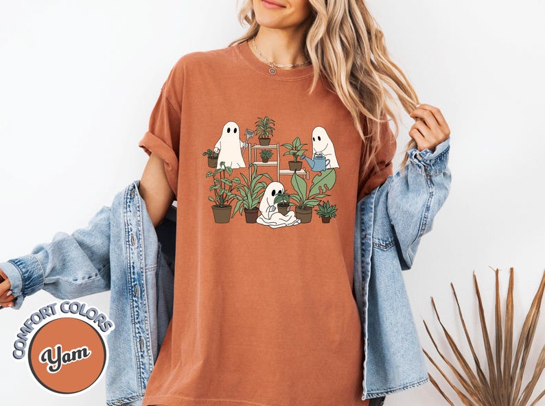 May include: Terracotta-colored t-shirt featuring a graphic of three cartoon ghosts tending to potted plants. The ghosts are white with black eyes, and the plants are green. The shirt has the words "COMFORT COLORS" and "Yam" on it.