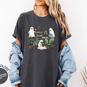 May include: Dark gray t-shirt featuring a whimsical illustration of three cartoon ghosts tending to potted plants. The ghosts are depicted watering, planting, and caring for various green plants. The shirt has the text "COMFORT COLORS Pepper" on the bottom left.