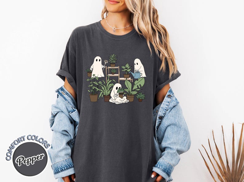 May include: Dark gray t-shirt featuring a whimsical illustration of three cartoon ghosts tending to potted plants. The ghosts are depicted watering, planting, and caring for various green plants. The shirt has the text "COMFORT COLORS Pepper" on the bottom left.