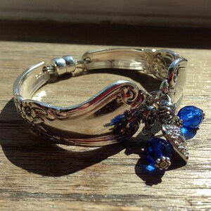 Pode incluir: Uma pulseira de prata feita de talheres reaproveitados. A pulseira apresenta um design floral e &eacute; adornada com contas e berloques de cristal azul. A pulseira &eacute; exibida em uma superf&iacute;cie de madeira.