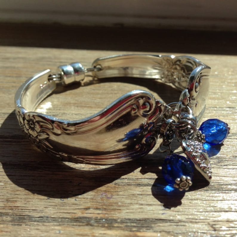 Pode incluir: Uma pulseira de prata feita de talheres reaproveitados. A pulseira apresenta um design floral e &eacute; adornada com contas e berloques de cristal azul. A pulseira &eacute; exibida em uma superf&iacute;cie de madeira.