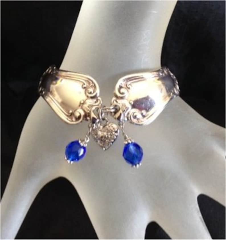 Pode incluir: Pulseira de prata feita com colheres, com cabe&ccedil;as de colher ornamentadas e um pingente em forma de cora&ccedil;&atilde;o. Duas contas azuis pendem do centro. A pulseira &eacute; mostrada em uma m&atilde;o.